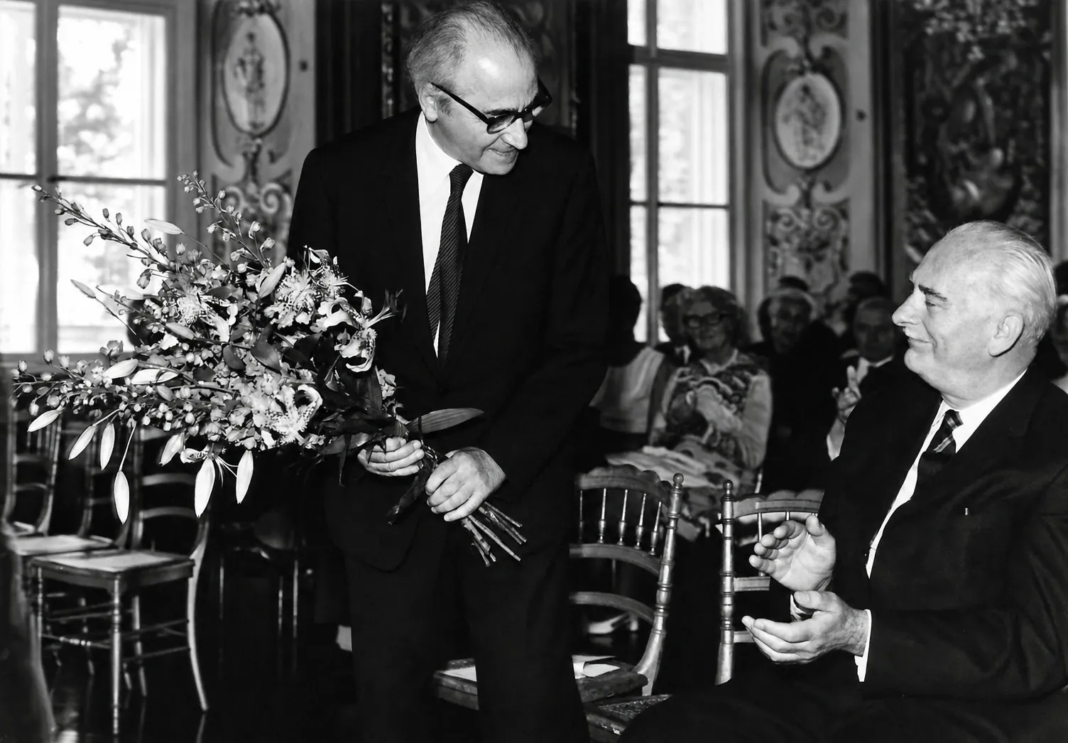 Otto Pächt mit einem Blumenstrauß in einem Festsaal, daneben ein applaudierender Herr